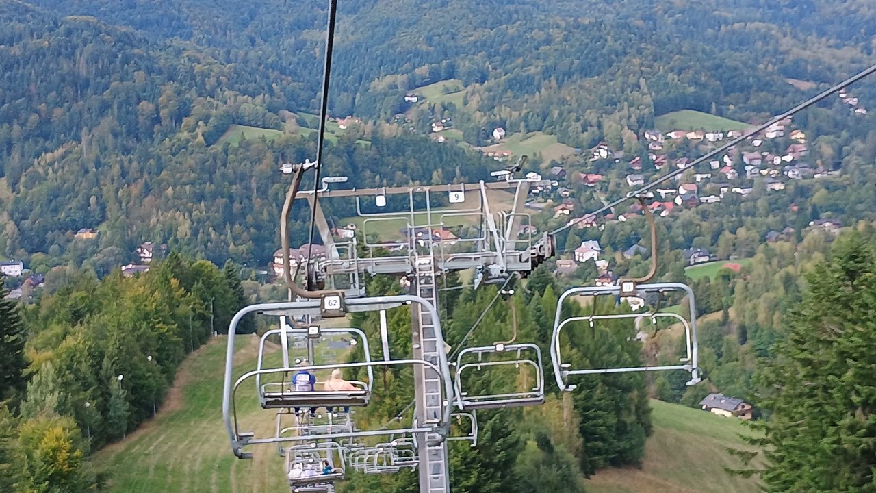 Widok z wyciągu krzesełkowego w górach na zalesione zbocza oraz panoramę miejscowości w dolinie.