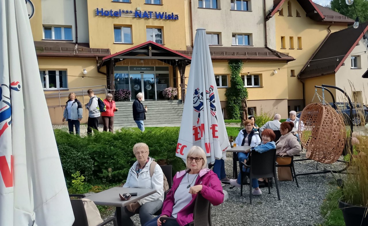 Grupa seniorów odpoczywająca na zewnątrz przed kompleksem Hotel NAT Wisła, przy stolikach z parasolami ogródka gastronomicznego.