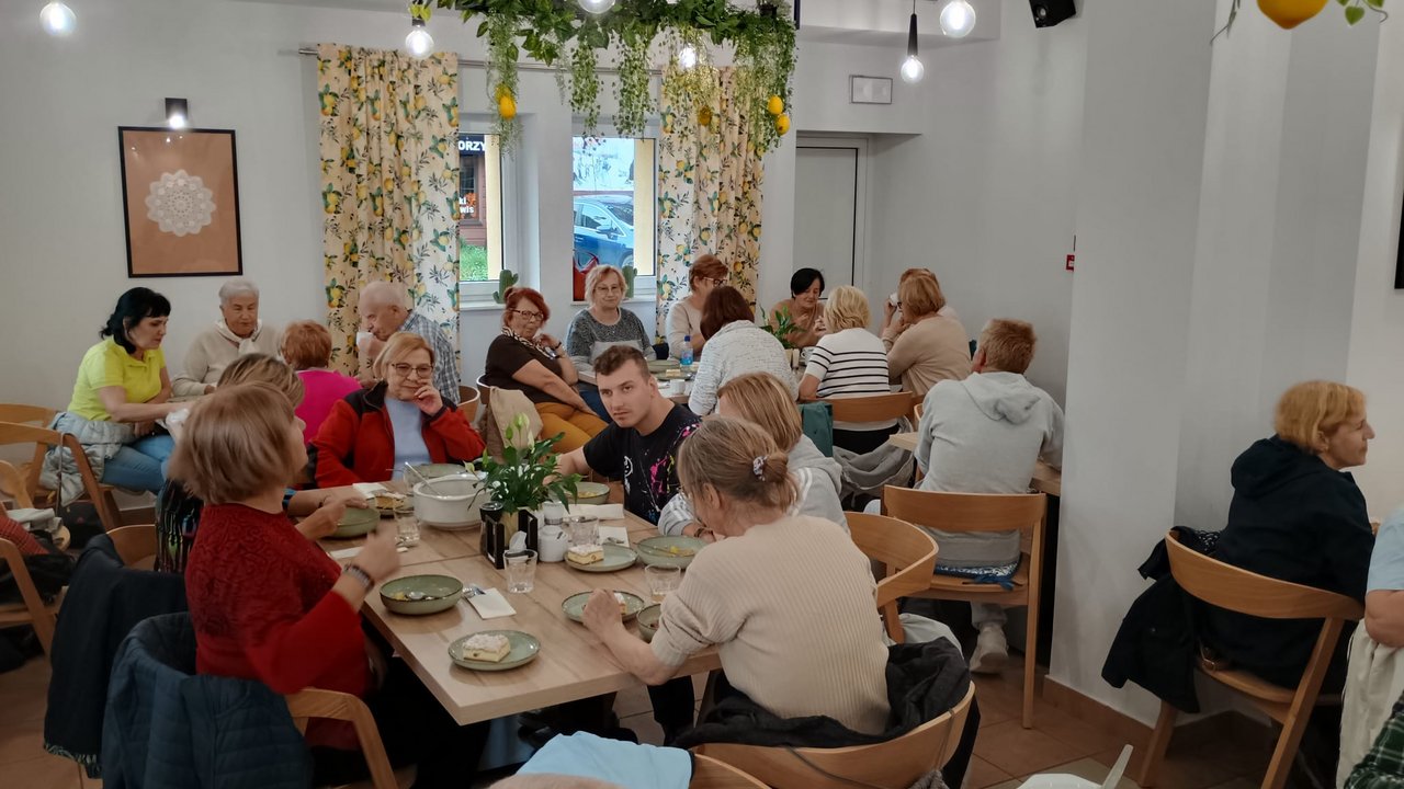 Grupa osób siedząca przy długim stole w jasnej, nowoczesnej restauracji ozdobionej roślinami, jedząca posiłek i rozmawiając