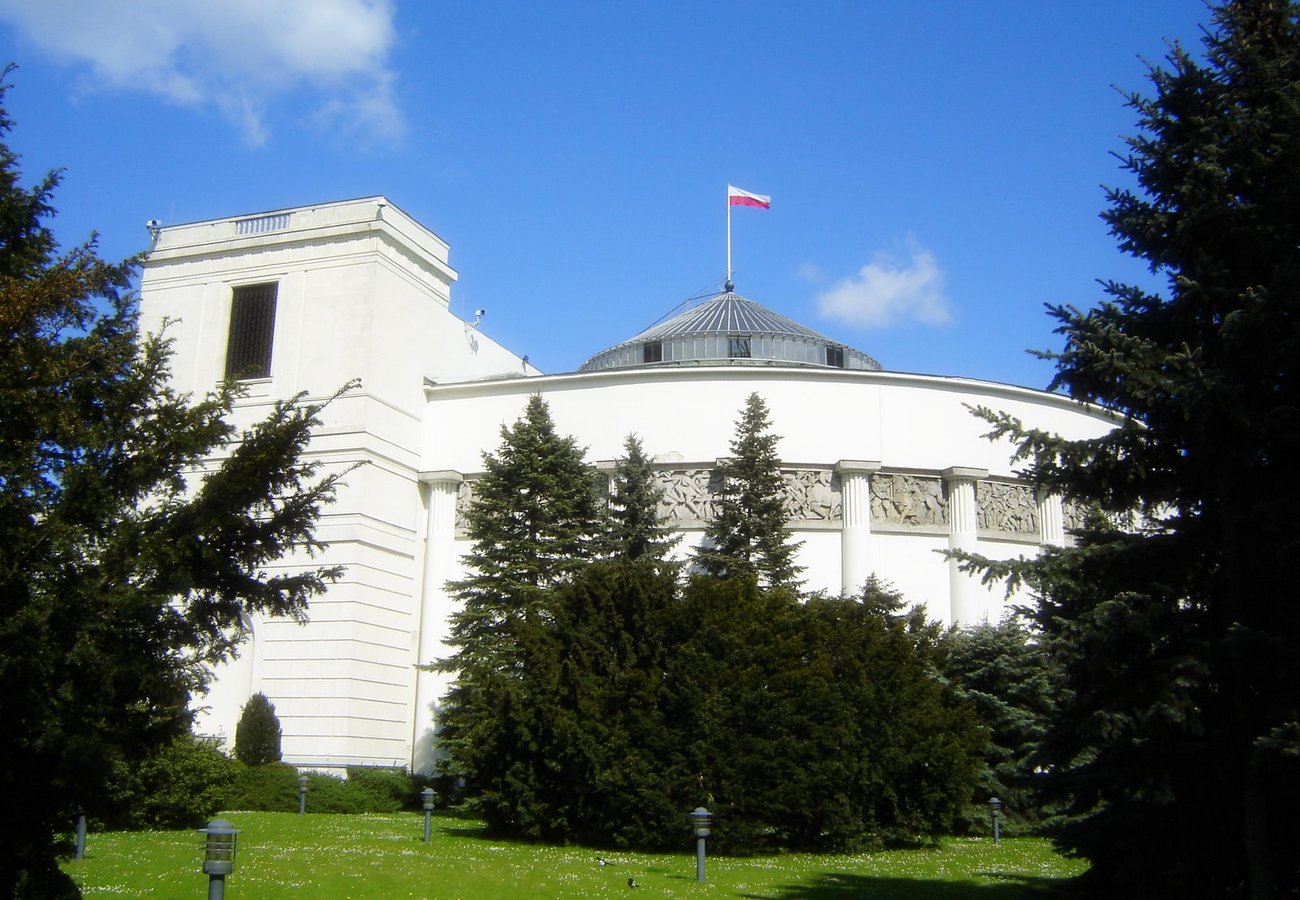  Biały gmach Sejmu RP z kopułą i polską flagą, otoczony zielenią, na tle błękitnego nieba